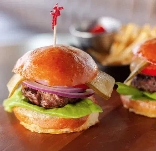 Two sliders with brioche buns, vegetables, and fries on wood.