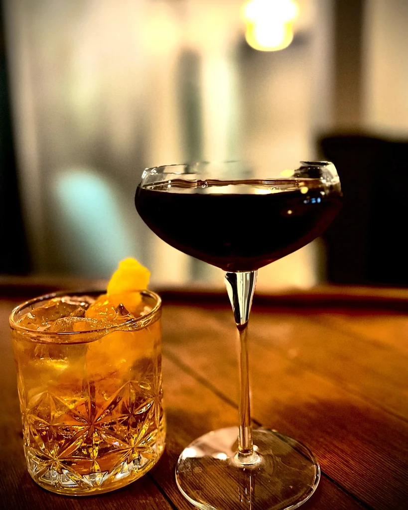 Two cocktails, one amber with orange peel, one dark, on a wooden surface.