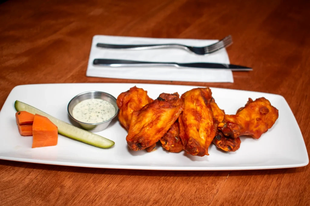 White plate of golden-brown chicken wings with red sauce, creamy dip, vegetables.