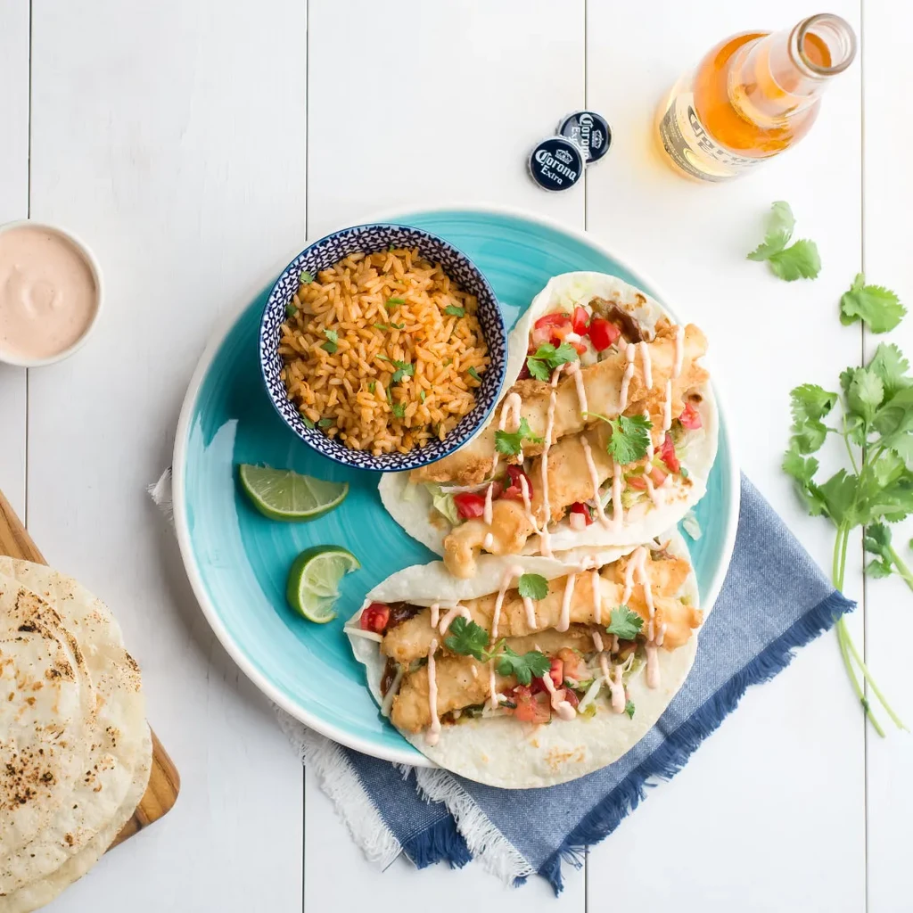 Overhead flat lay of fish tacos, rice, lime, sauce, and Corona beer.