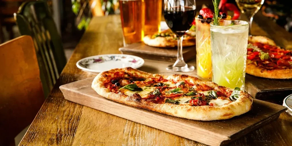 Two pizzas, beers, wine, and cocktails on a wooden restaurant table.