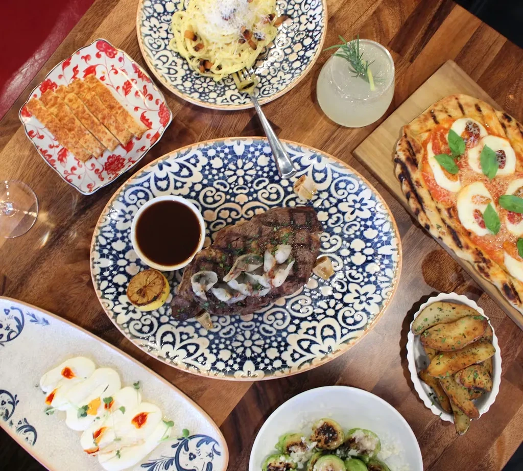 Flat lay of diverse restaurant dishes: steak, pasta, pizza, and drink.