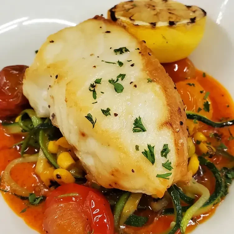 Pan-seared cod with vegetables and sauce, garnished with parsley and lemon.