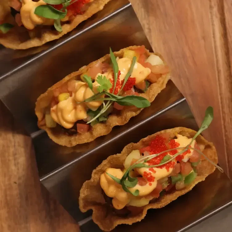 Three tacos in wavy shells with toppings, sauce, and microgreens.