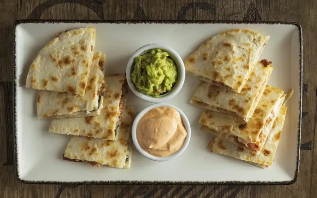Quesadilla wedges with guacamole and orange dip on a wooden table.