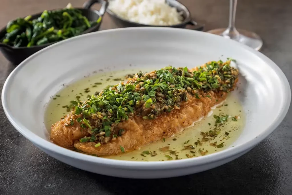 Pan-seared fish with green herbs, sauce, and sides in a white bowl.