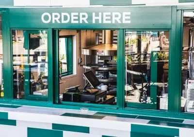 Green fast-food service window, 'ORDER HERE' text, checkered counter, kitchen.