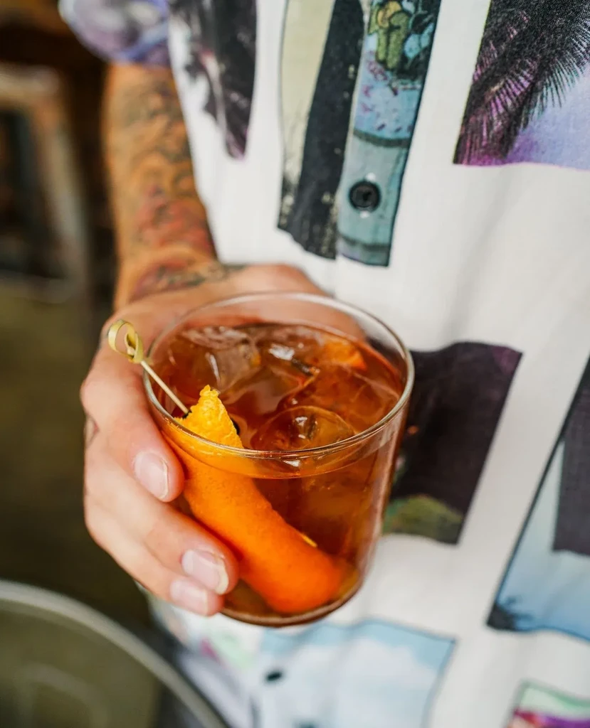 Person holding an amber cocktail with ice, orange peel, and citrus garnish.