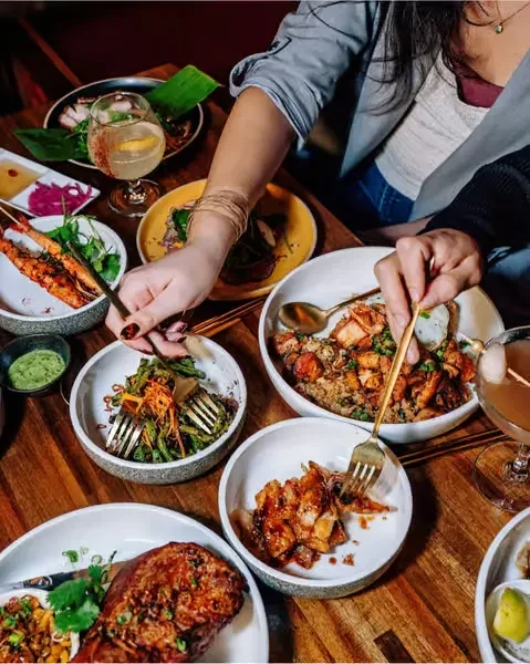 People share a meal and cocktails at a wooden table.