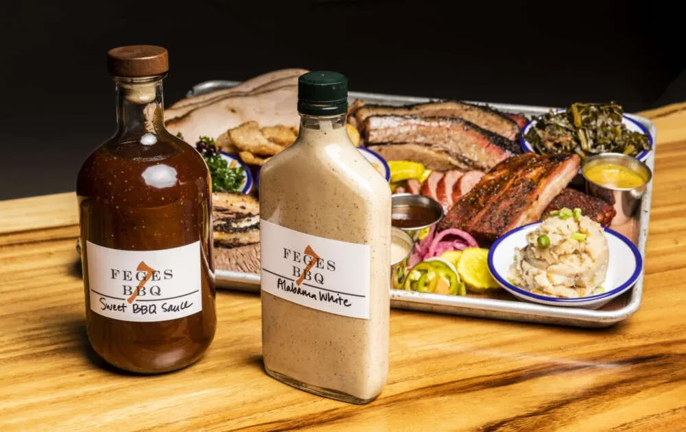 Two Feges BBQ sauces, brisket, ribs, and sides on a wooden table.