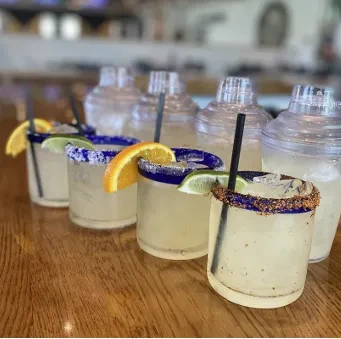 Five margarita drinks with blue rims and citrus garnishes on a bar.