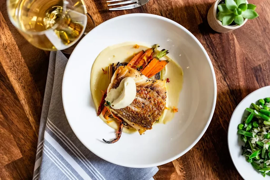 Pan-seared fish, roasted vegetables, and sauce in a white bowl.