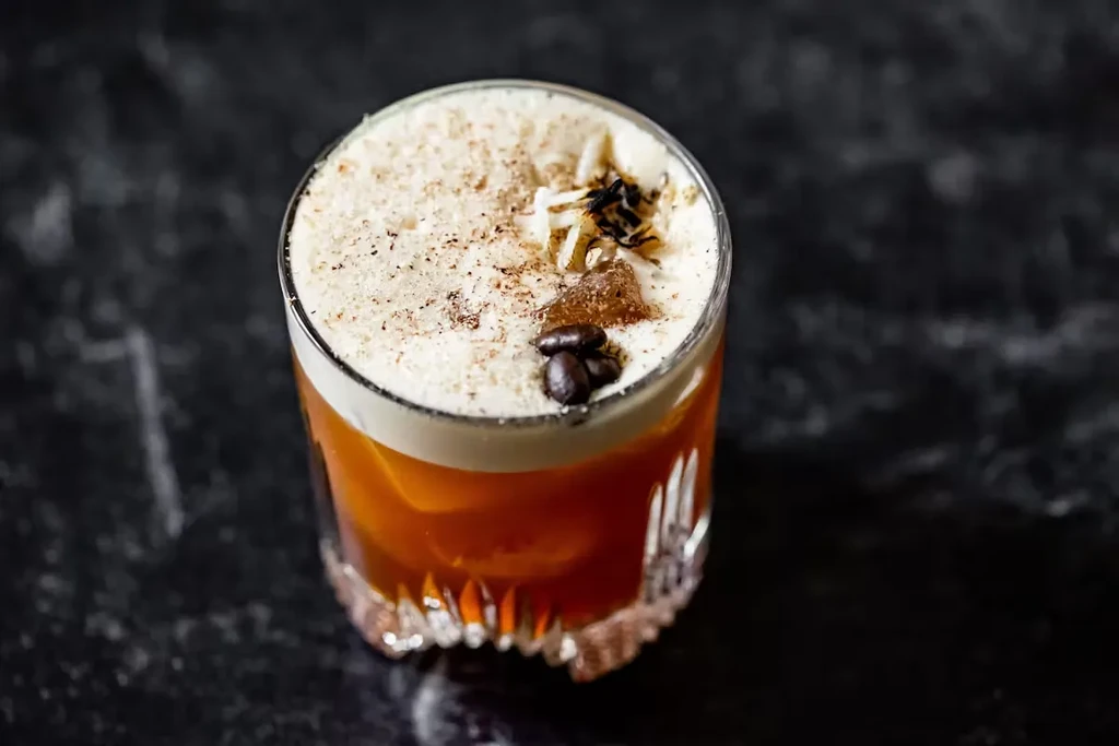 Cocktail with amber liquid, white foam, spices, and coffee beans.