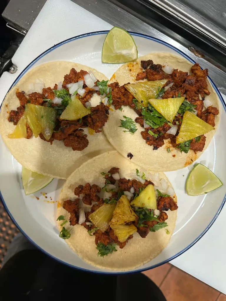 Three al pastor tacos garnished with pineapple, onions, cilantro, and lime.