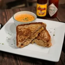 Grilled cheese sandwich, tomato soup, and hot sauces on a dark table.
