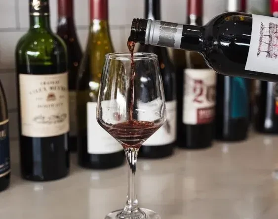 Red wine pours into a glass, with blurred bottles in background.