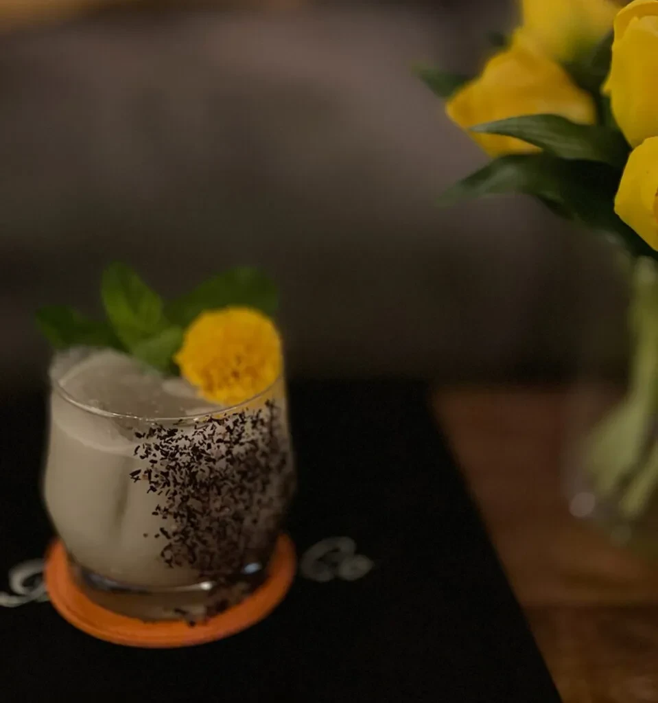 Cocktail in short glass with mint, yellow flower, and dark sprinkles.