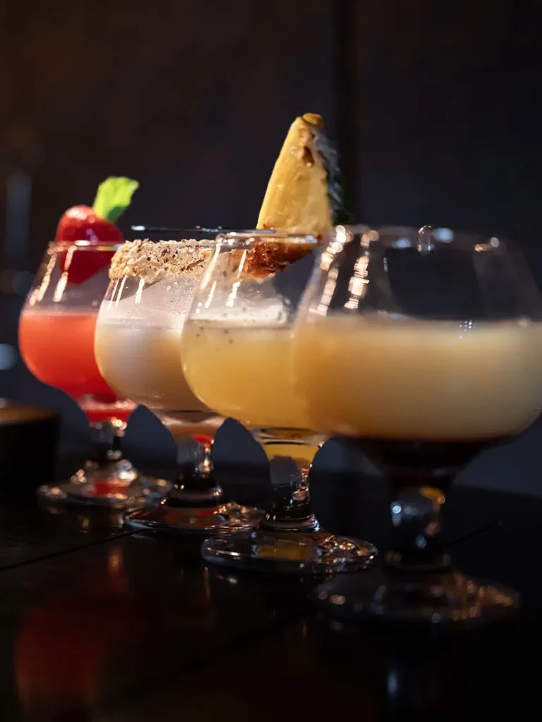Four elaborate cocktails in stemmed glasses on a dark, reflective surface.