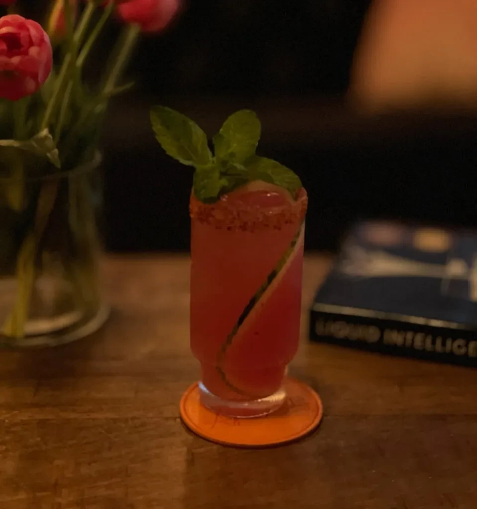 Red-pink cocktail with mint, cucumber, and spiced rim on wooden table.