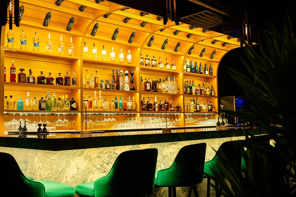 Well-stocked bar with glowing shelves, dark counter, and green bar stools.