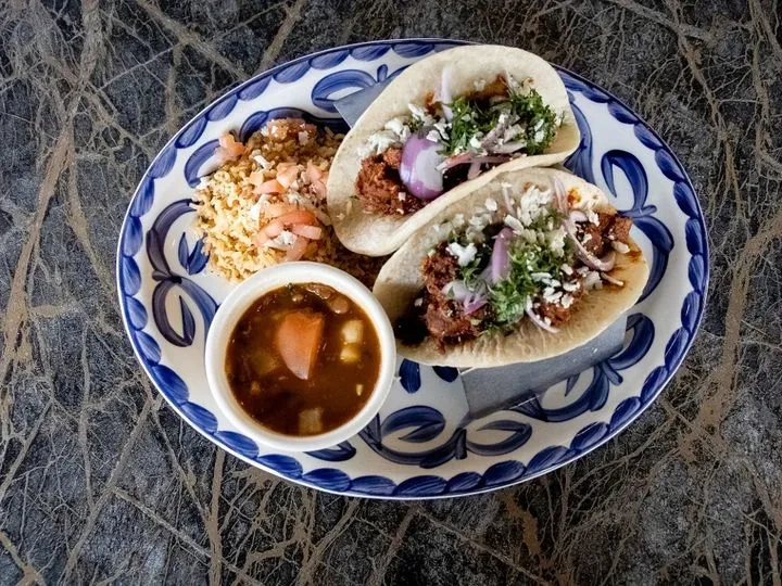 Overhead shot of two tacos, rice, and soup on a decorative plate.
