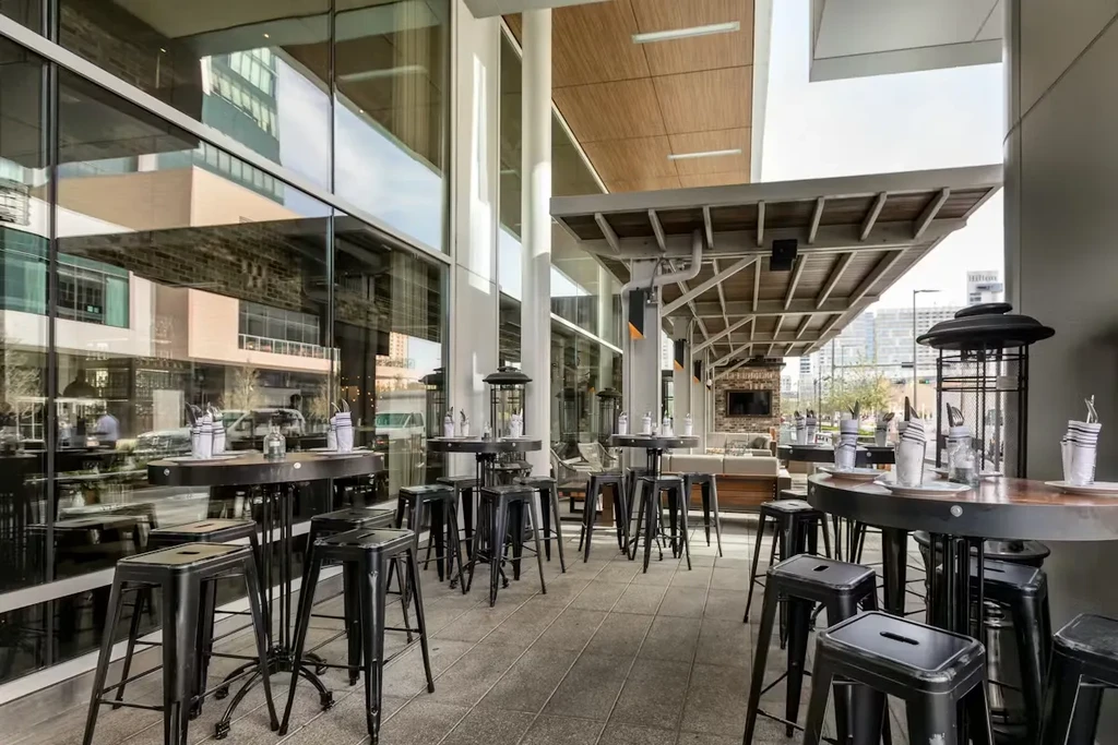 Outdoor restaurant patio with high-top tables, modern ceiling, and glass facade.