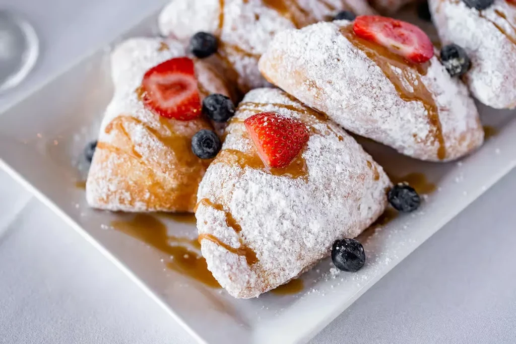 Fried dough pastries with powdered sugar, syrup, strawberries, and blueberries.