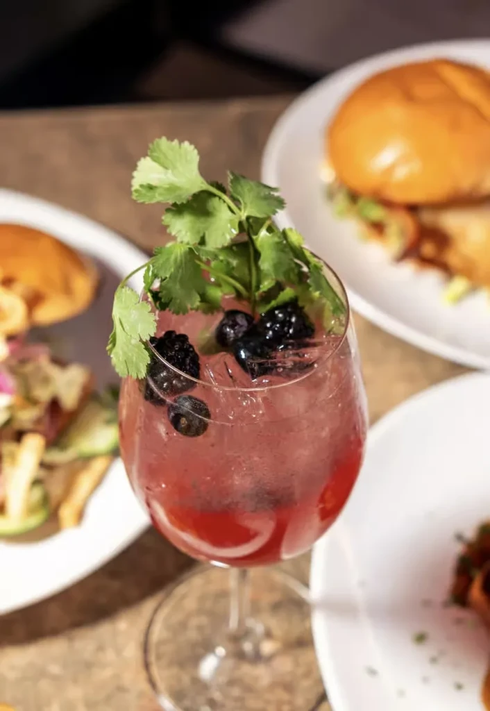 Red cocktail in wine glass with berries and cilantro.