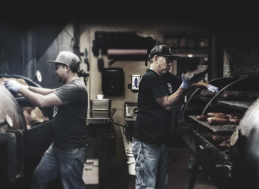 Two men in gloves place food in large barbecue smokers.