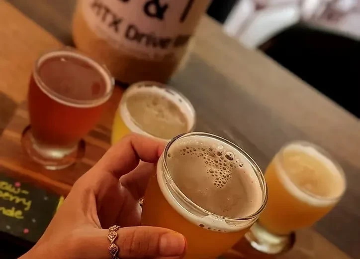 Hand holding a glass of beer from a flight on a wooden paddle.