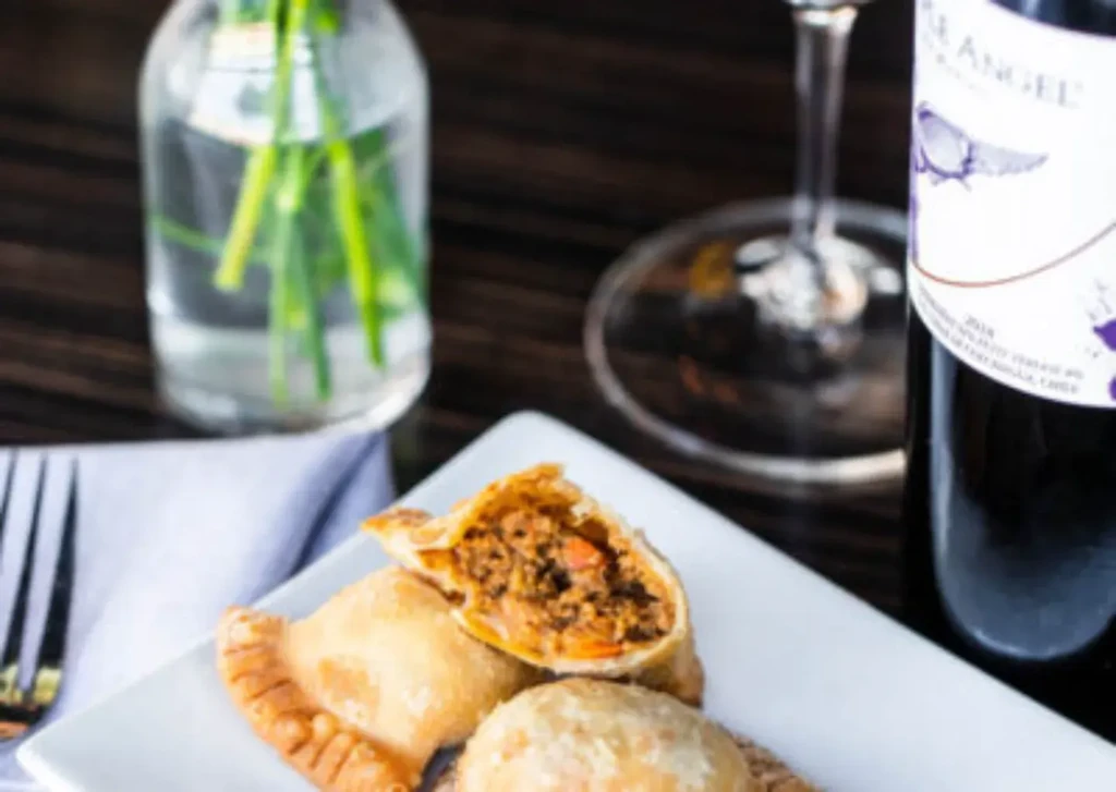 Plate of golden pastries filled with meat, wine bottle in background.