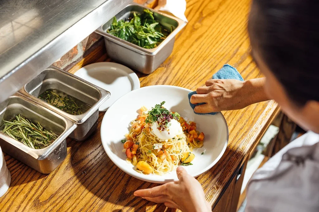 Chef plates pasta dish with herbs and vegetables in kitchen.