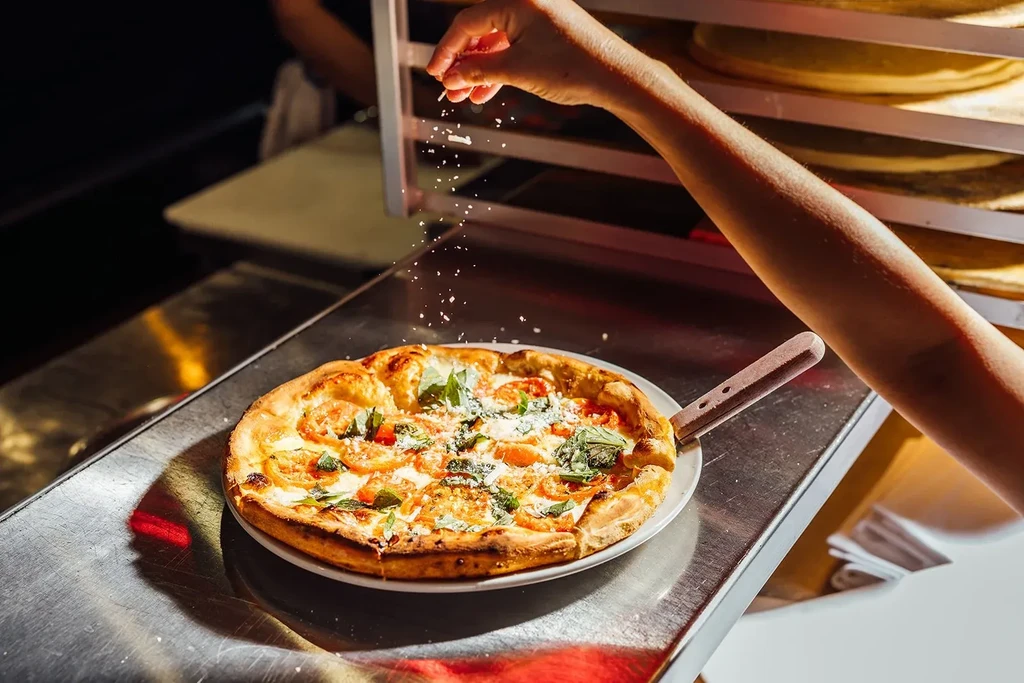 Hand sprinkles cheese over pizza with tomatoes and basil.