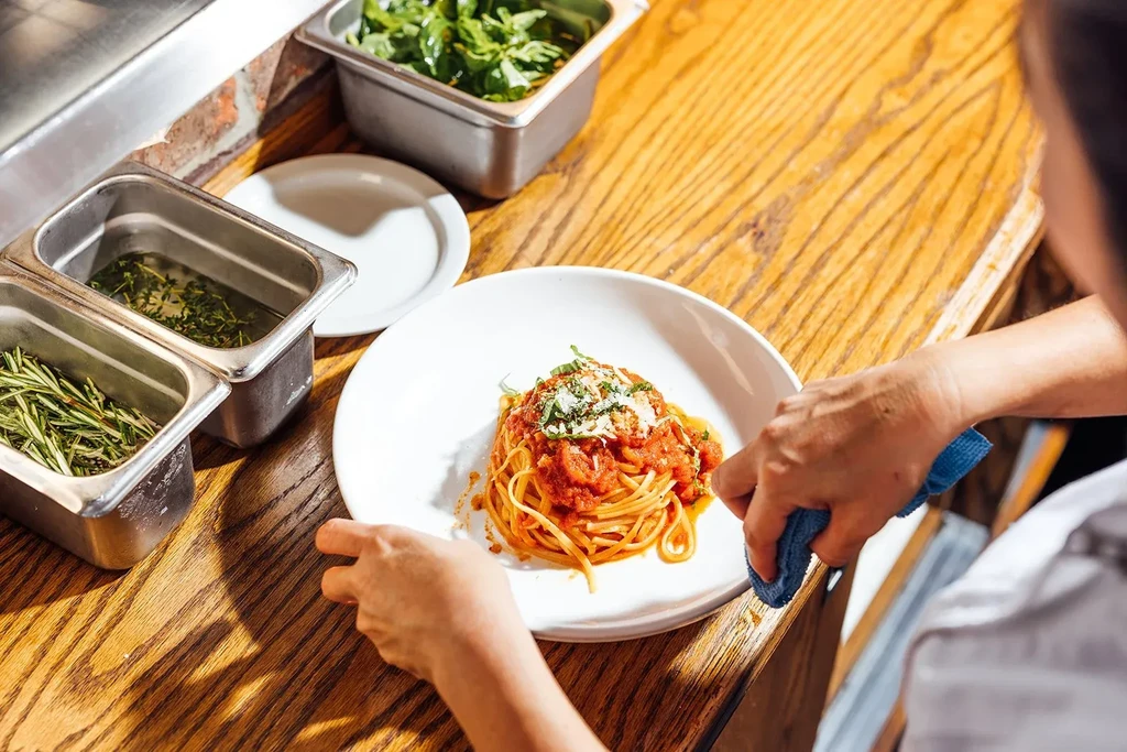 Chef's hands adjust a plate of spaghetti with sauce and herbs.