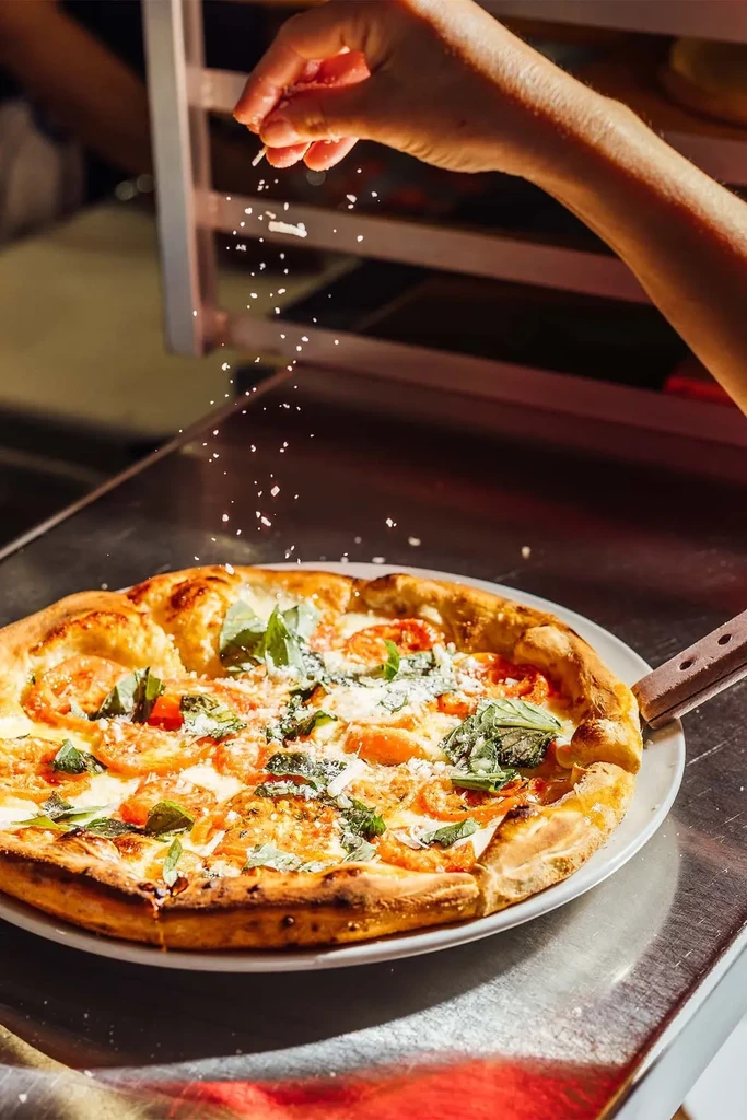 A person sprinkles flakes over a pizza with cheese, tomatoes, and basil.