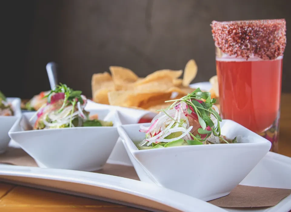 Ceviche appetizers, michelada, and chips arranged on a wooden table.