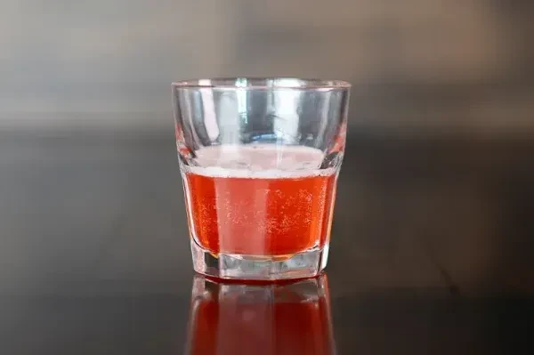 Fizzy reddish-orange beverage in a clear glass on a dark surface.