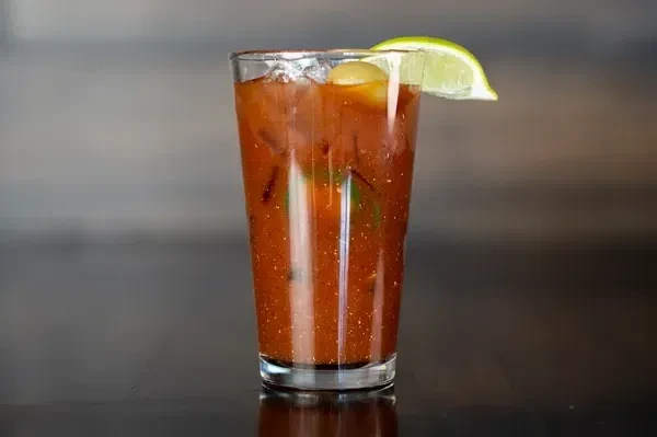 Glass of reddish-brown beverage with lime, olive, and ice.