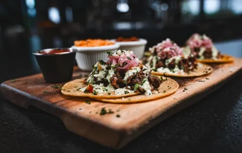Three tacos with meat, sauce, herbs, and pickled onions on a board.