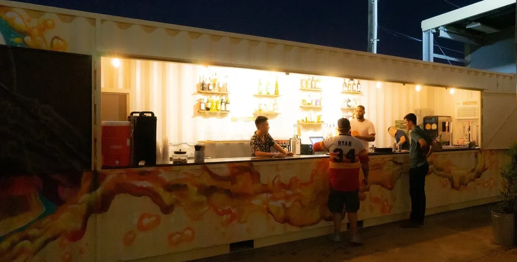 Four men at an outdoor container bar with bottles and murals.