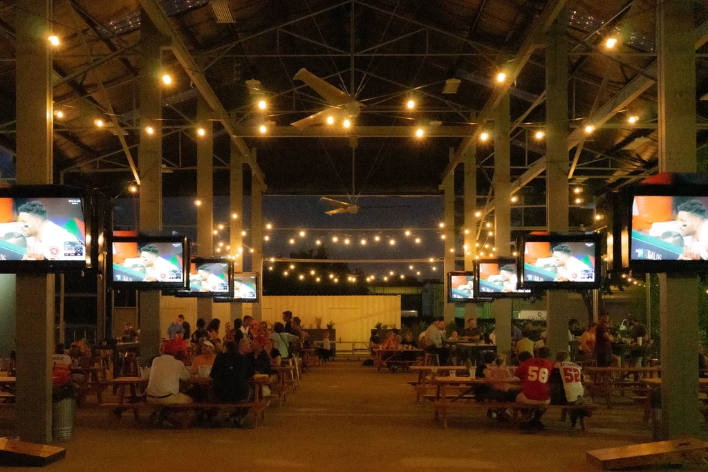 Open-air restaurant/bar with string lights, TVs showing sports, and patrons.