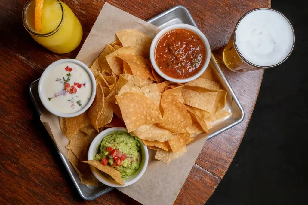Overhead view: tortilla chips, salsa, queso, guacamole, juice, and beer.