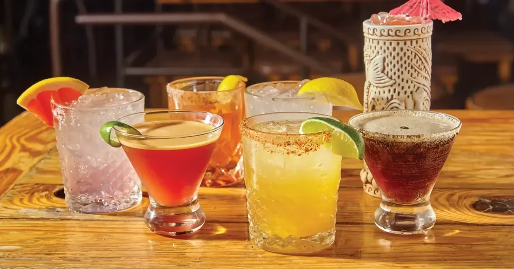 Seven different cocktails with garnishes are arranged on a wooden table.