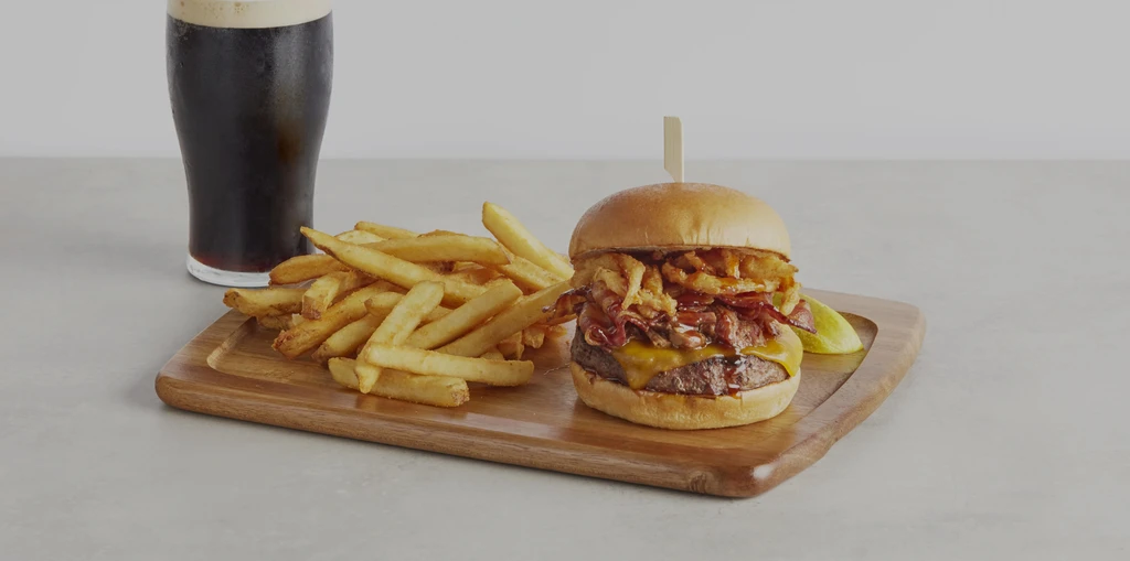 Gourmet burger, fries, and cold dark drink on a wooden board.