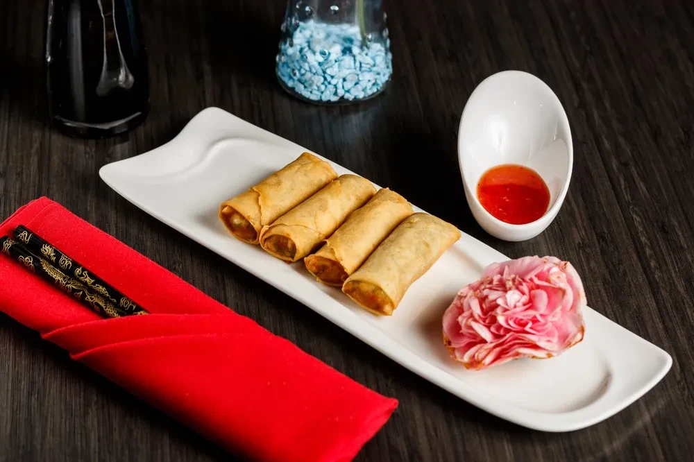 Four spring rolls, pink flower, red sauce, and chopsticks on a plate.