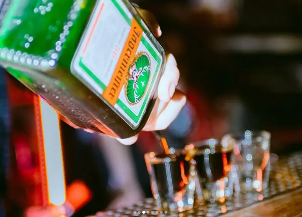 Hand pouring Jägermeister from a bottle into shot glasses.