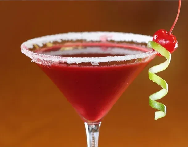 Red cocktail in a martini glass with cherry and lime peel.