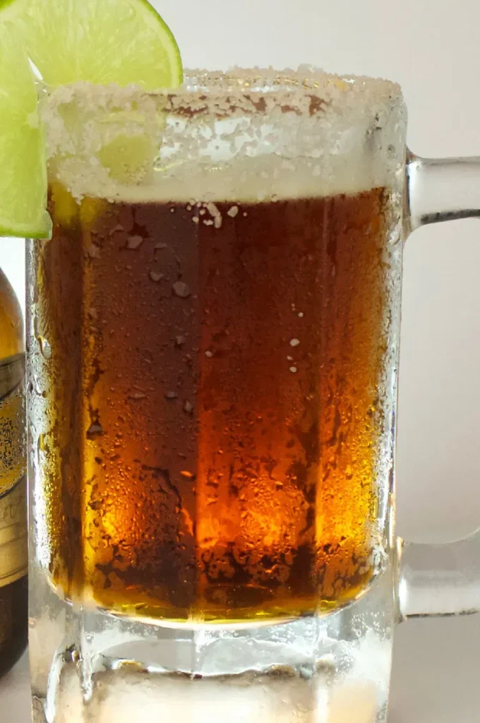 Salt-rimmed glass mug with dark amber drink and lime slices.