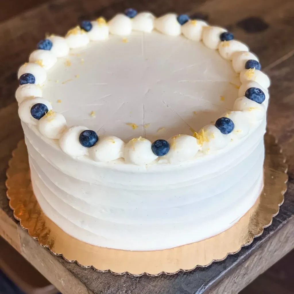 Round white-frosted cake with blueberries, lemon zest, and ridged sides.
