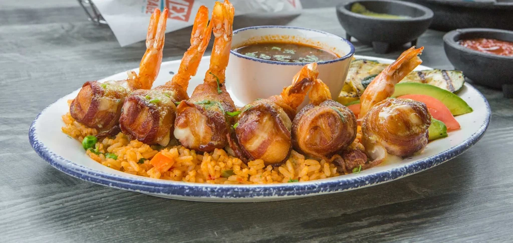 Bacon-wrapped shrimp, rice, vegetables, and sauce on a white plate.